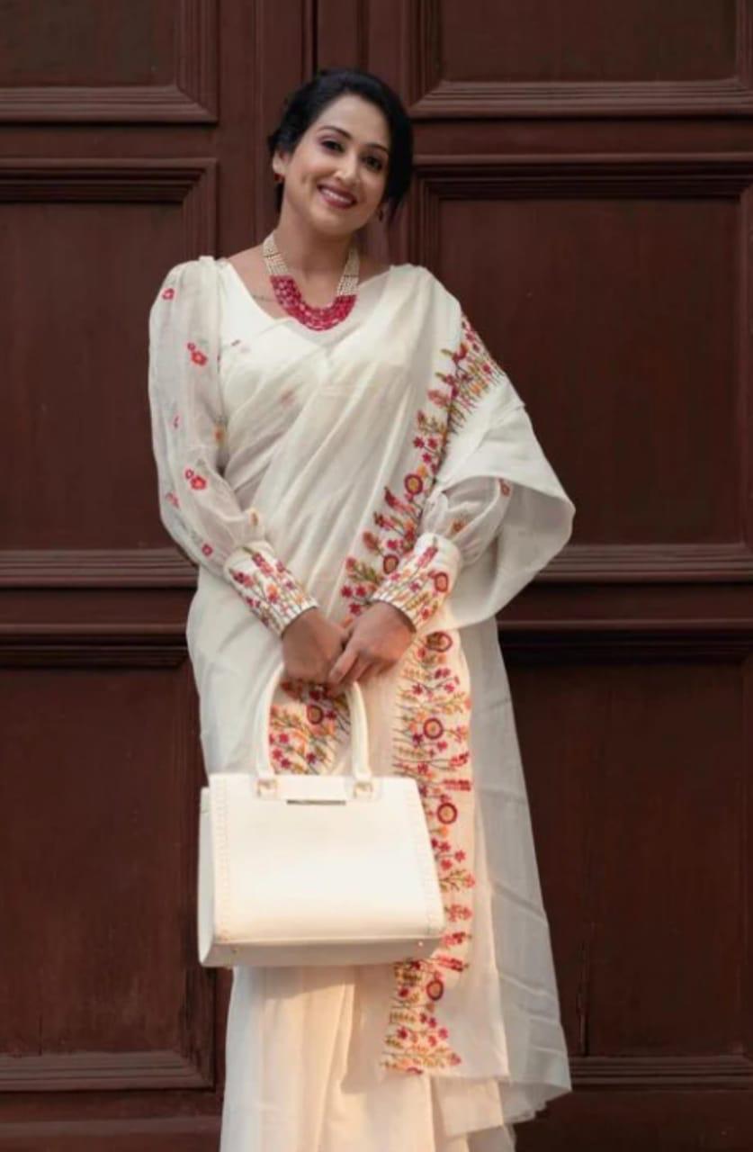 Woman standing in white cotton saree with  an ivory cotton saree with red and orange floral embroidery on the border and handbag